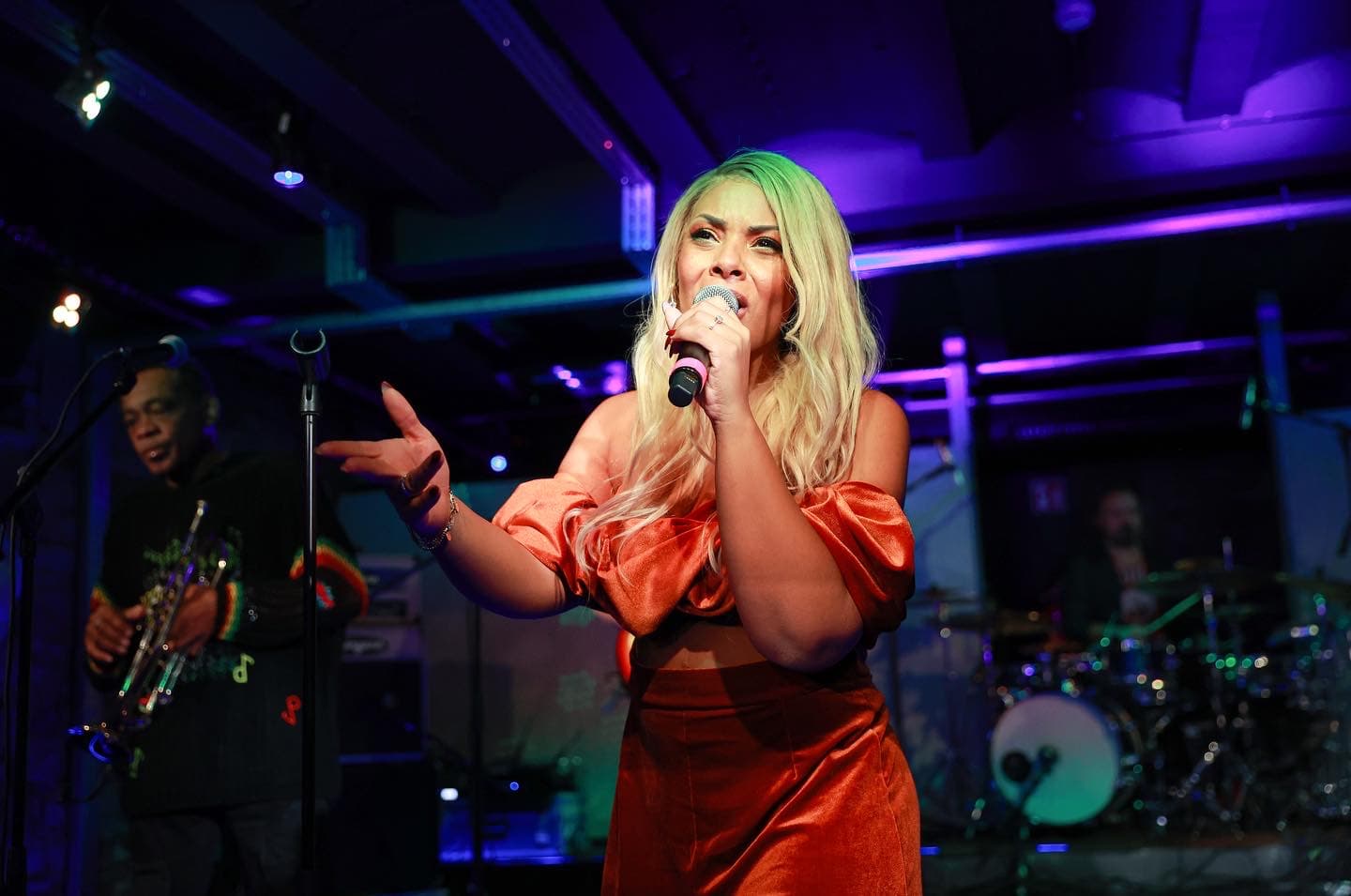 Amy Sue im orangefarbenen Satin-Outfit singt mit Saxophonist bei blauem Licht