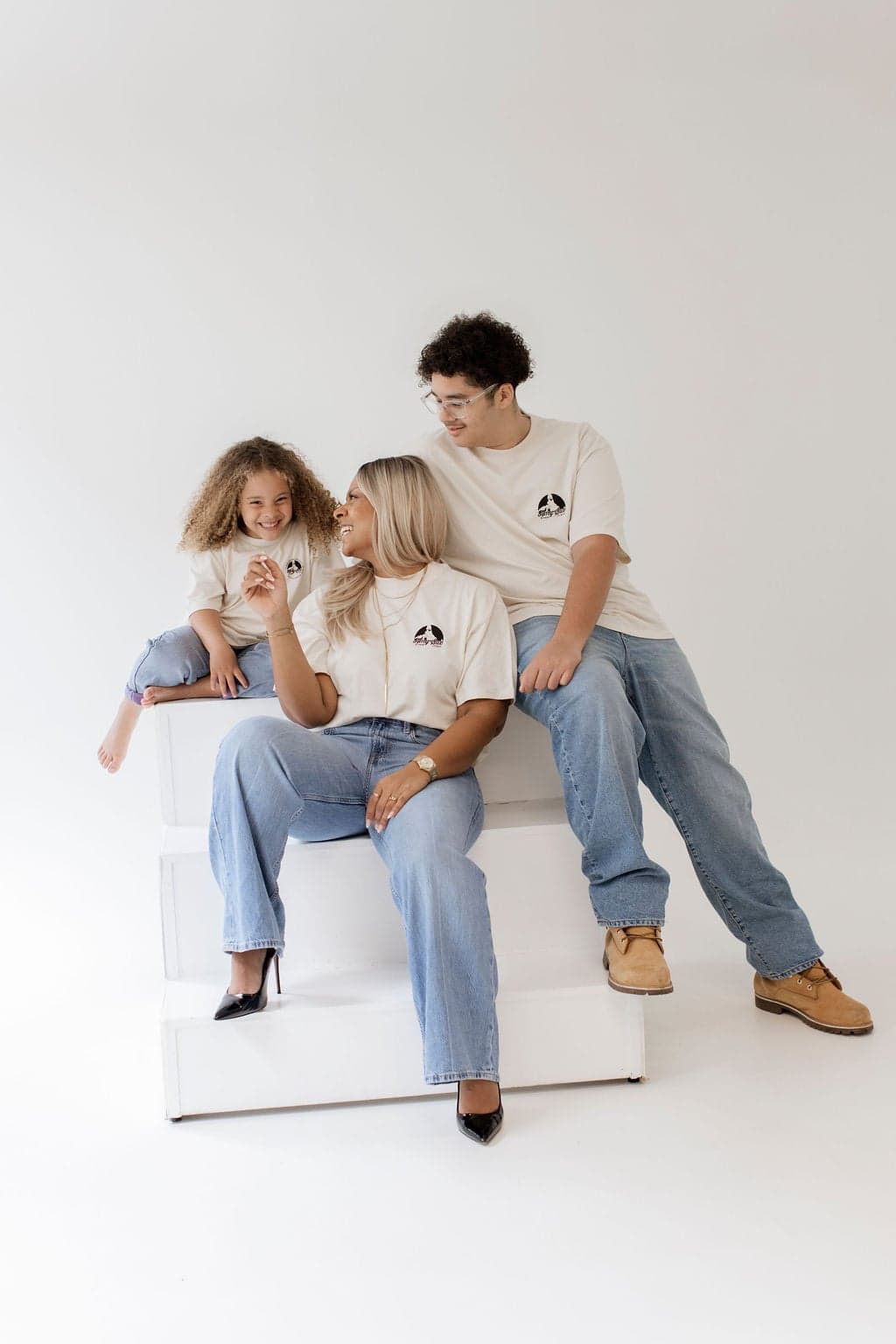 Amy Sue sitzt mit einem Kind und einem Jugendlichen auf weißen Stufen, alle drei in der Jubiläums-Kollektion, entspannt lachend