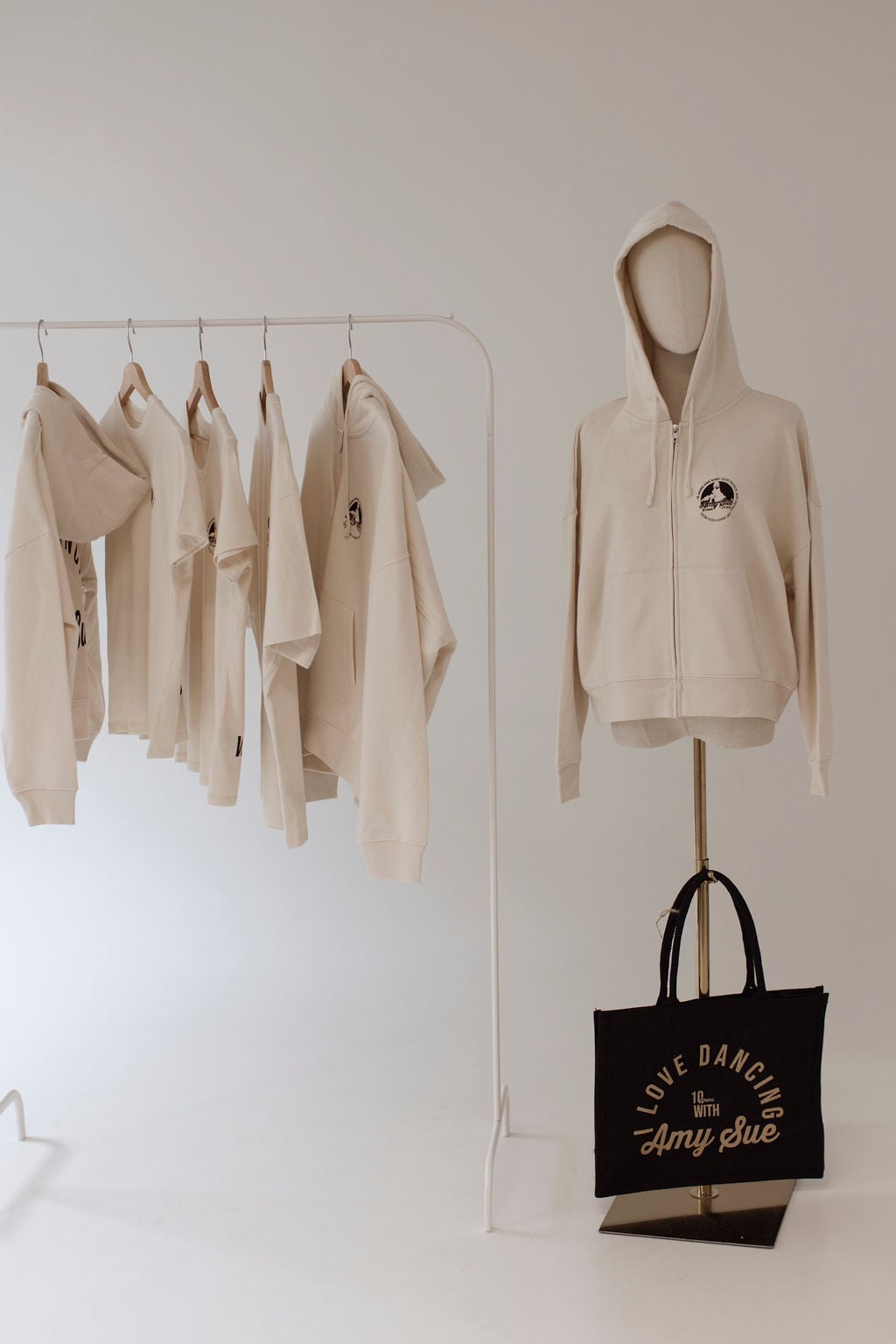 Die komplette Jubiläums-Kollektion im Studio: Shirts und Hoodies auf der Kleiderstange links, Zipper-Hoodie auf der Schaufensterpuppe rechts und die schwarze Beachbag mit Gold-Logo prominent unten rechts am Brass-Stand