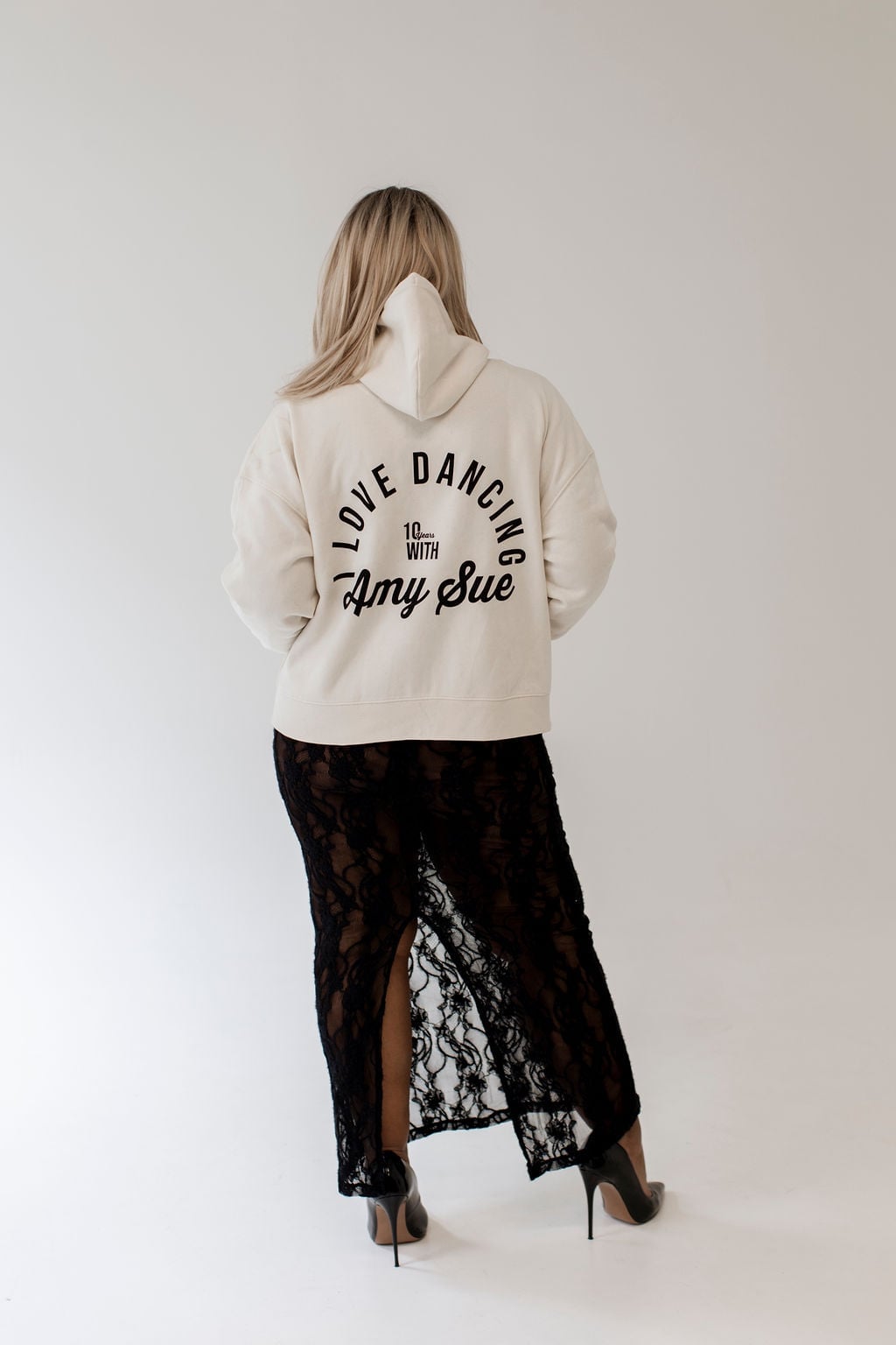 Frau mit blonden Haaren von hinten im Cream Pullover Hoodie — runder Backprint „I LOVE DANCING WITH Amy Sue 10 Years" voll sichtbar, schwarzer Spitzen-Maxirock und Stilettos, Editorial-Statement