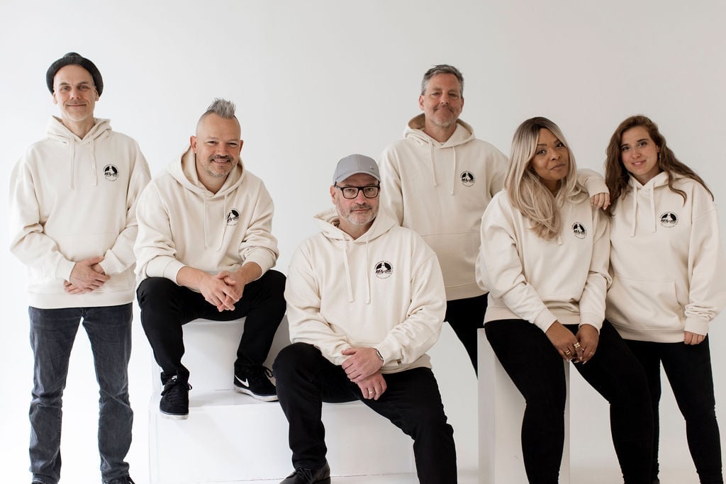 Amy Sue und sechs Bandmitglieder, alle im cremefarbenen Pullover Hoodie — drei sitzend, vier stehend, entspanntes Ensemble-Portrait vor weißem Studio-Hintergrund