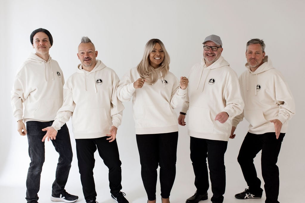 Line-up: Amy Sue mit vier Bandmitgliedern, alle stehend im cremefarbenen Pullover Hoodie, lachend und entspannt posiert — dynamische Community-Aufnahme