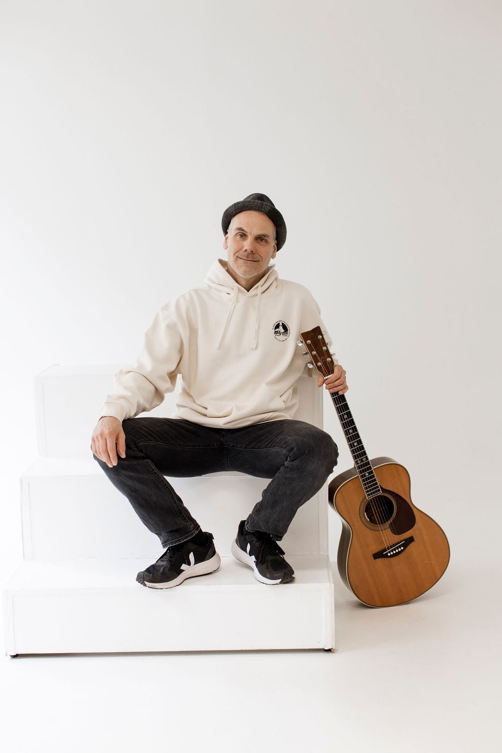 Bandmitglied sitzend auf einer weißen Stufe im cremefarbenen Pullover Hoodie mit Chest-Logo, Akustikgitarre neben sich angelehnt — editorial Studio-Portrait