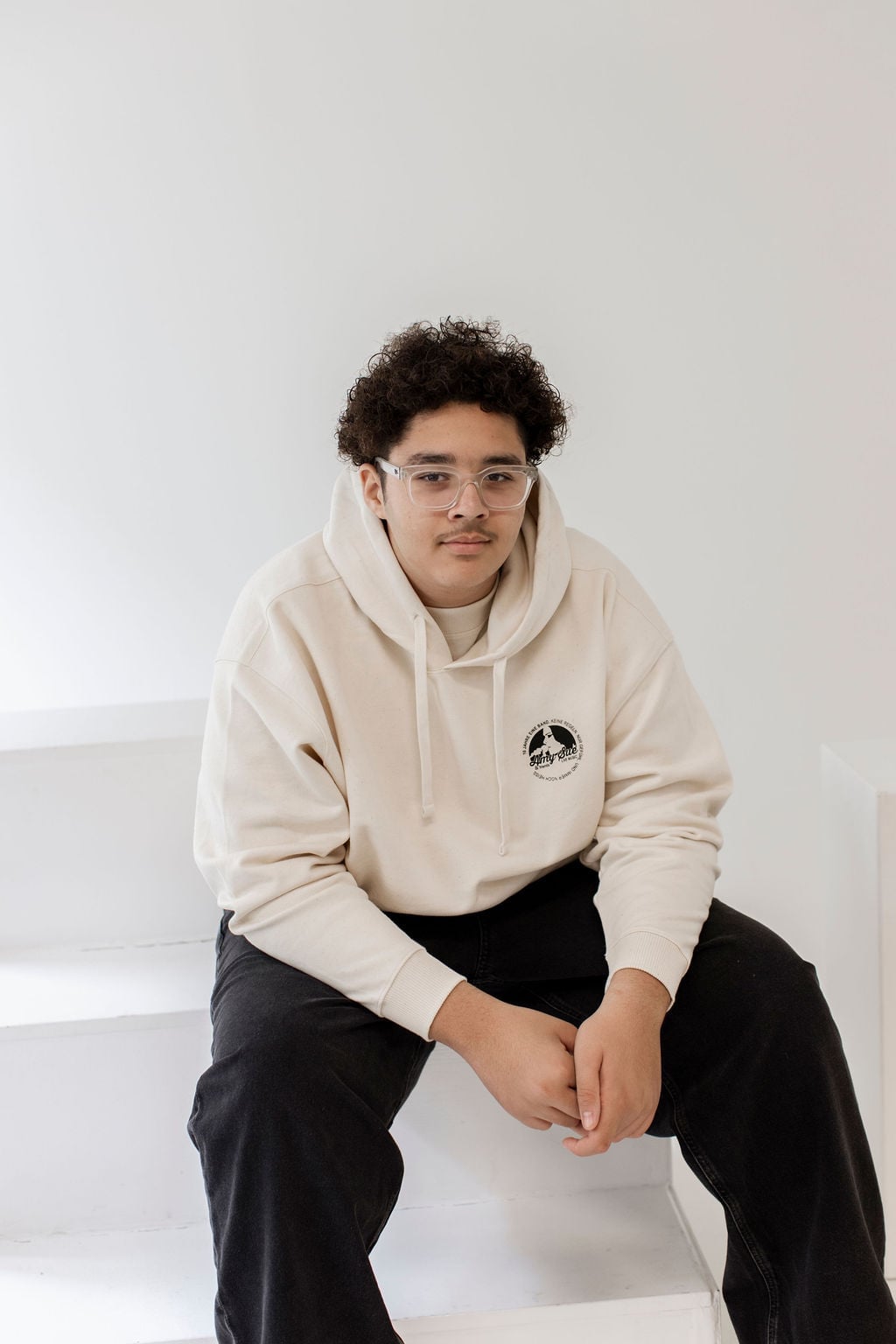 Jüngeres Bandmitglied sitzend im Pullover Hoodie mit Chest-Logo-Stempel („10 Jahre eine Band, keine Regeln"), ruhiges Editorial-Portrait — nächste Generation in der Jubiläums-Kollektion