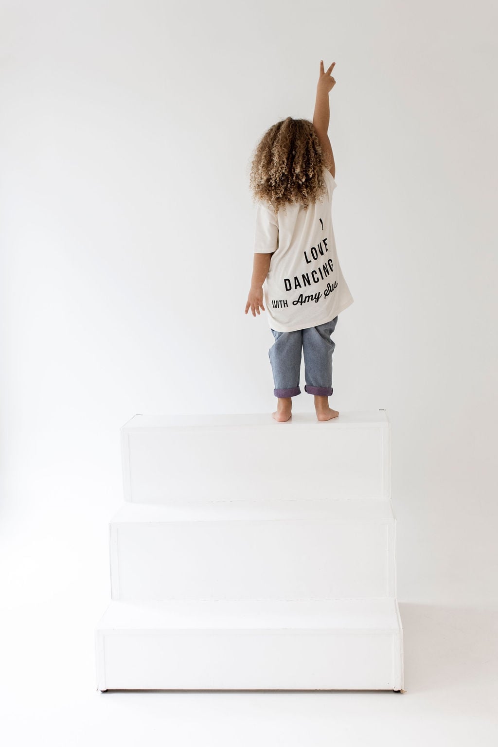 Kind mit Curly Hair von hinten auf weißen Stufen im Cream Kindershirt — vollständiger „I LOVE DANCING WITH AMY SUE"-Backprint sichtbar, Hand mit Peace-Zeichen erhoben