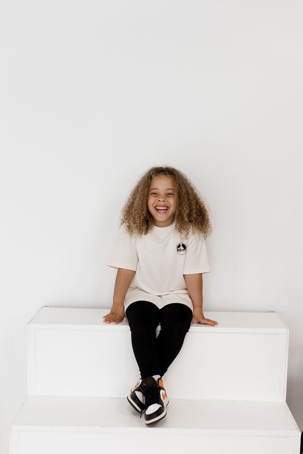 Lachendes Kind mit Curly Hair sitzt auf weißen Stufen im Cream Kindershirt mit Chest-Logo, schwarze Leggings und orangefarbene Sneakers — lebendiges Studio-Editorial