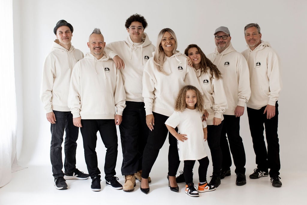 Gruppenportrait von Amy Sue mit ihren sieben Band- und Familienmitgliedern: alle in cremefarbenen Pullover-Hoodies mit Chest-Logo, ein Kind vorne im Kindershirt, lachend frontal in die Kamera — die komplette Community der Jubiläums-Kollektion