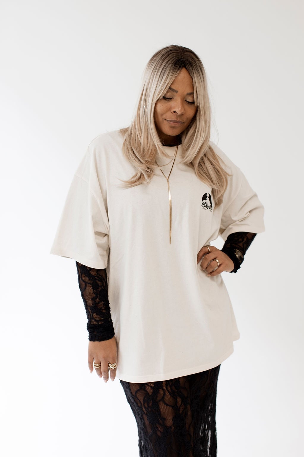 Amy Sue im Cream Oversized T-Shirt mit schwarzen Spitzen-Sleeves darunter — sanfter Blick nach unten, Y-Goldkette, Chest-Logo sichtbar, intimes Studio-Editorial