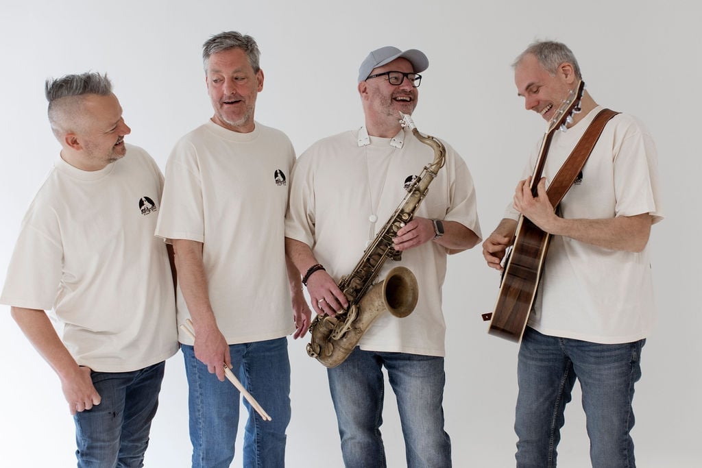 Vier Bandmitglieder mit Instrumenten in den T-Shirts — Drumsticks, Saxophon und Gitarre, gemeinsam lachend