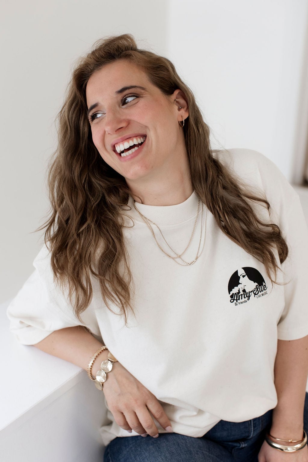 Brünette Frau im Oversize T-Shirt, entspannt an weißer Wand lehnend, lachend